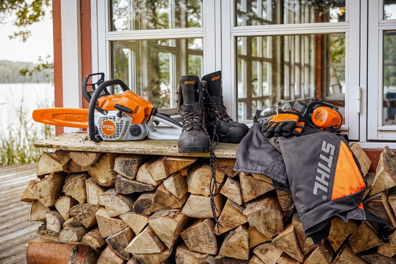 Îmbrăcăminte de protecție STIHL și MS 162 pe un raft, deasupra lemnelor de foc stivuite