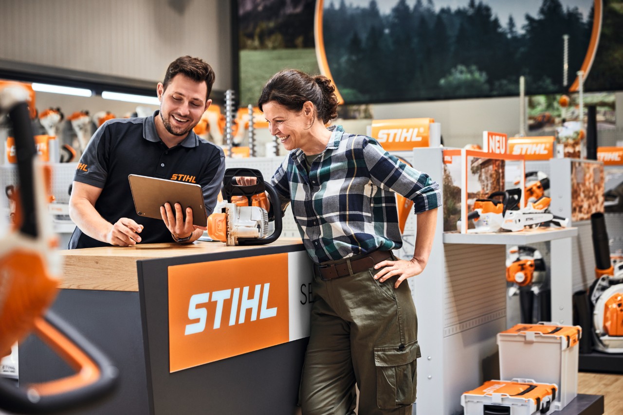 Un distribuitor STIHL consiliază o tânără la cumpărarea unei foarfeci de tuns garduri vii STIHL în fața unui raft de vânzări