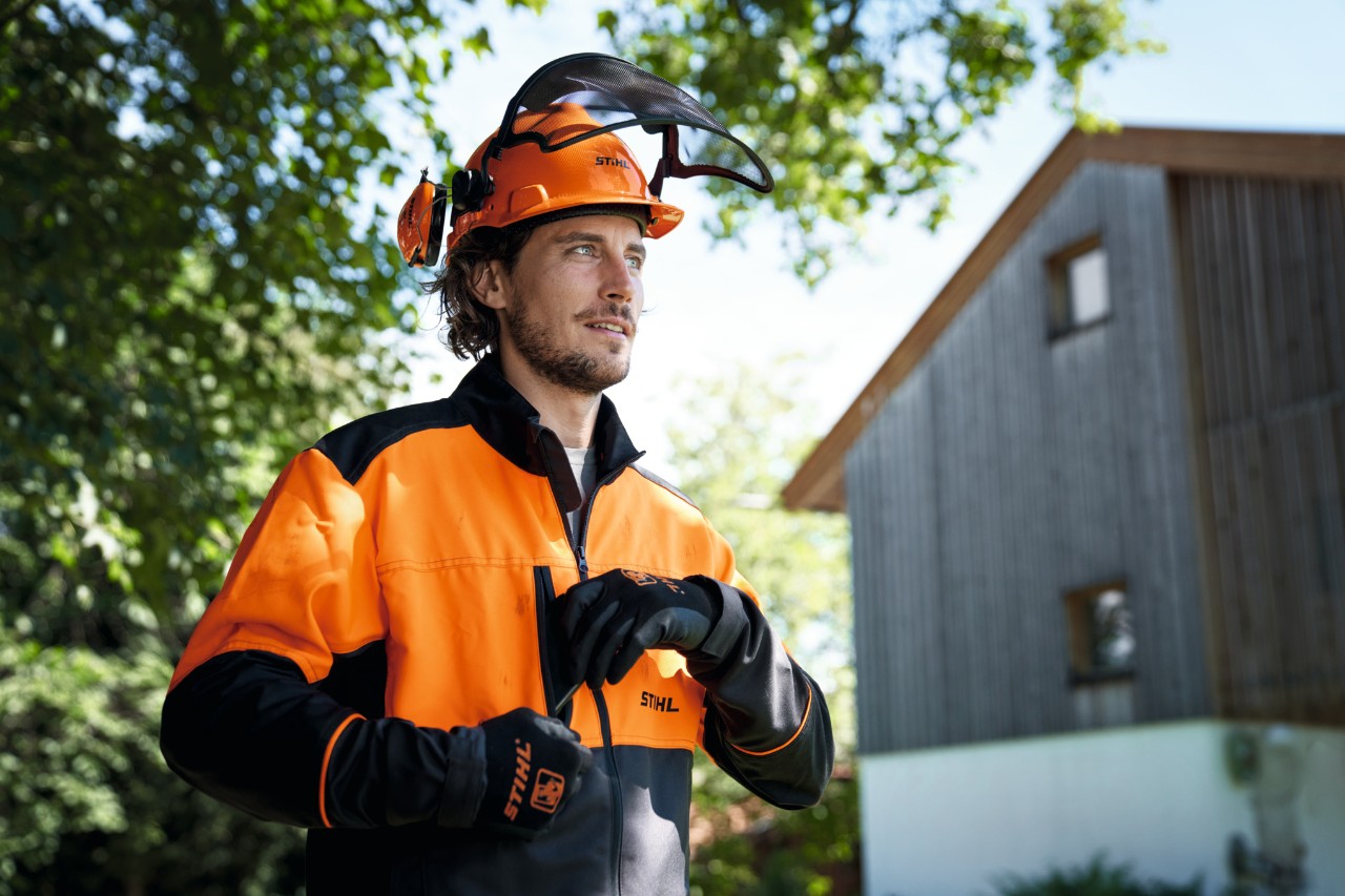 Un bărbat care poartă echipament de protecție STIHL portocaliu și negru, inclusiv cască și mănuși, privește încrezător în afara imaginii, cu o casă și copaci în fundal 