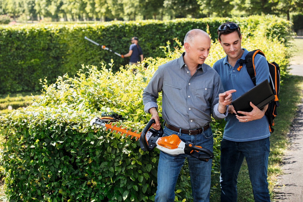 Dr. Nikolas Stihl ține în mână un trimmer de tuns garduri vii STIHL și vorbește cu o persoană care se află lângă el și care ține un laptop.