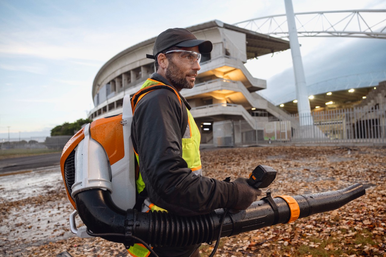 Spre seară, o persoană curăță frunzele și resturile de pe un stadion folosind suflanta fără fir ALLPRO STIHL BRA 600.