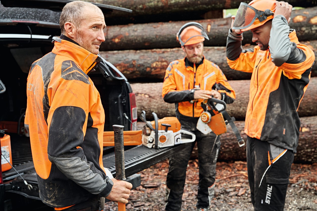 Trei bărbați în echipament de protecție STIHL și cu utilaje cu acumulator STIHL lângă portbagajul deschis al unui vehicul, cu o stivă de lemne în fundal