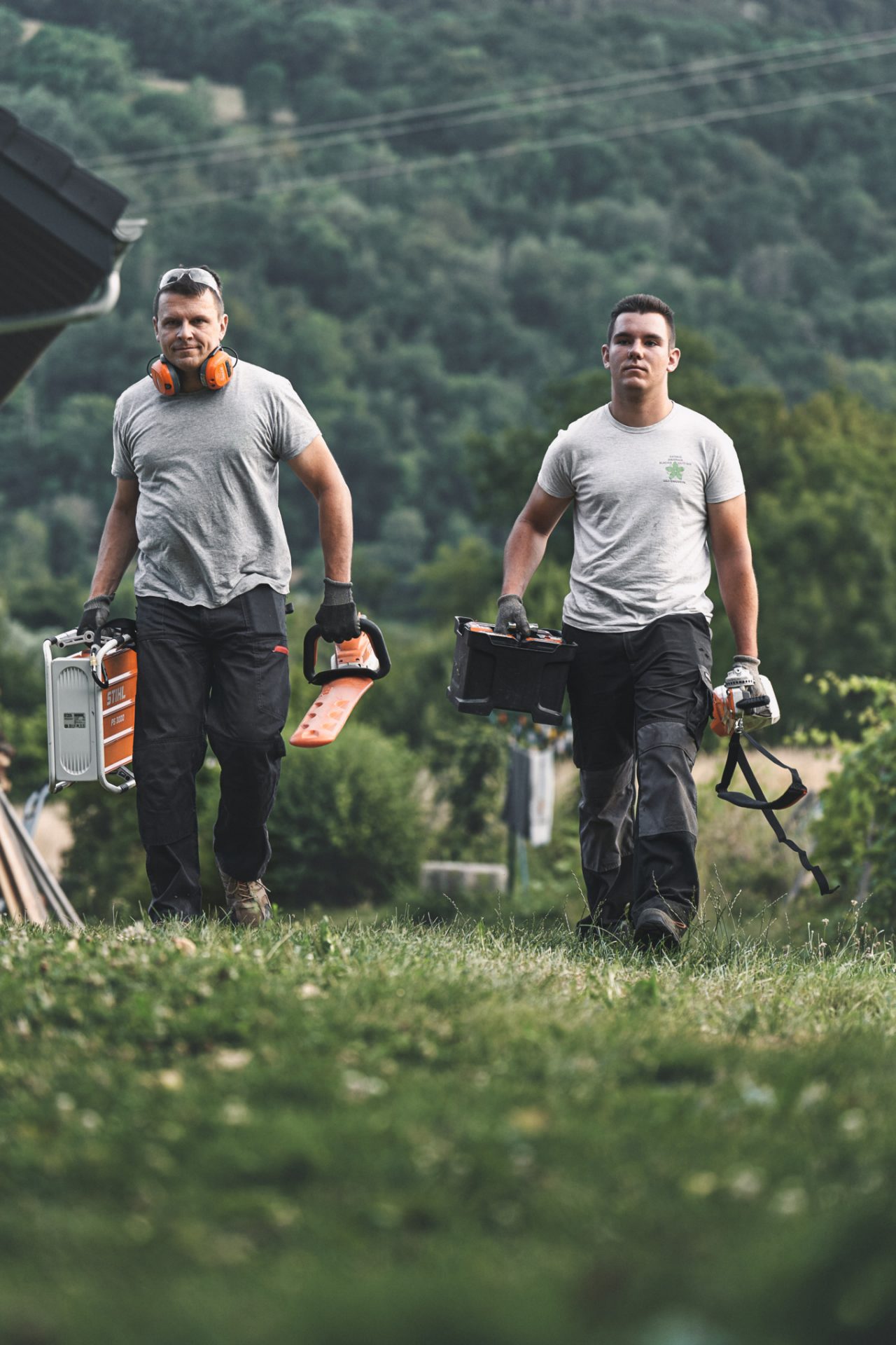 Doi profesioniști merg cu utilajele cu acumulator STIHL pe o pajiște