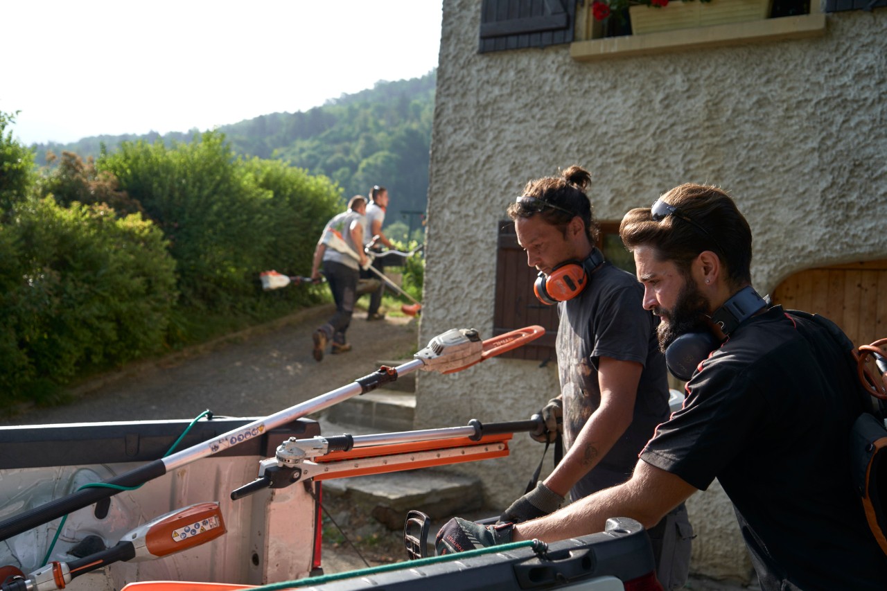 Doi peisagiști descarcă utilajele cu acumulator STIHL dintr-o mașină, cu alți lucrători în fundal 