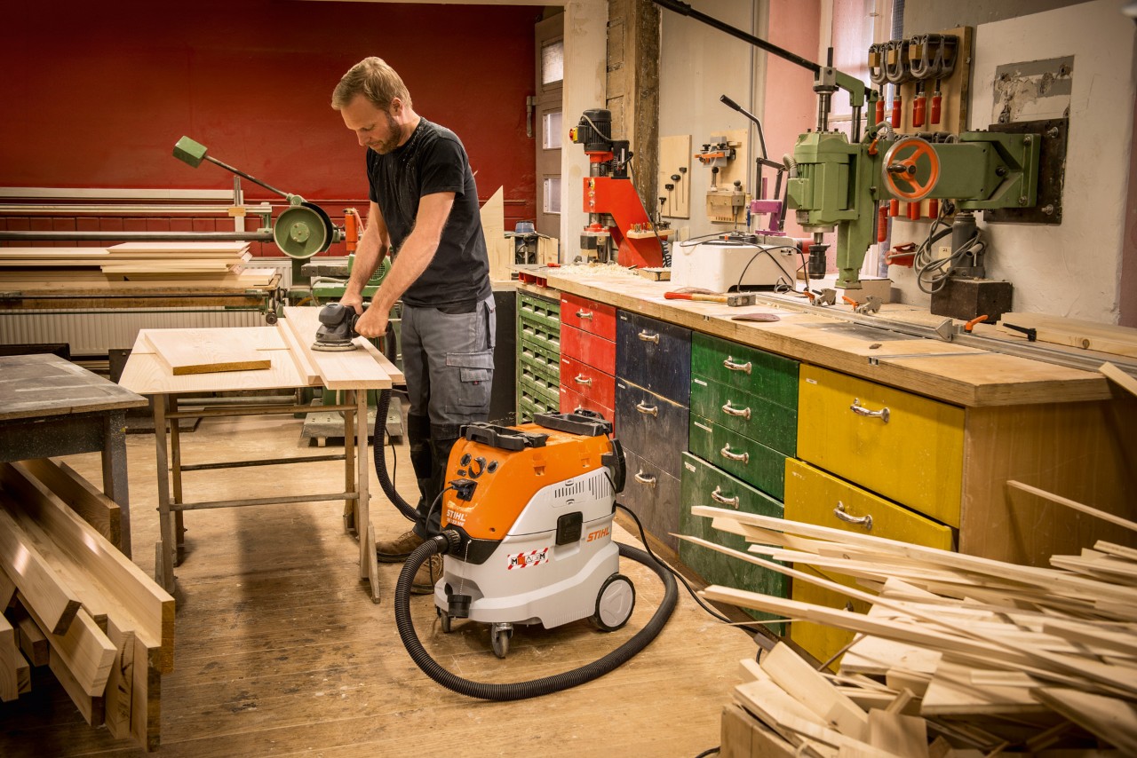 Un bărbat utilizează aspiratorul pentru suprafețe umede și uscate STIHL SE 133 ME într-un atelier în timp ce lucrează cu scânduri din lemn