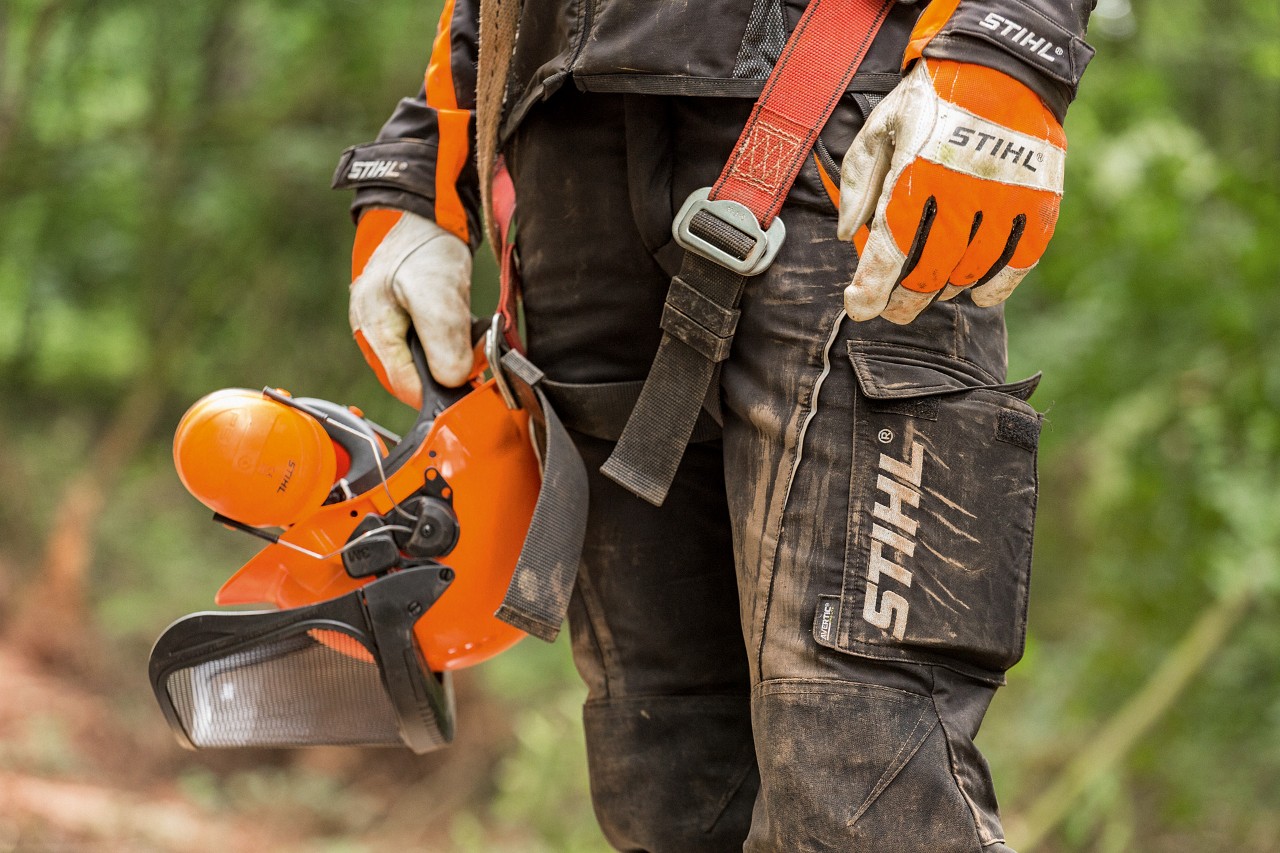 O persoană cu pantaloni de protecție împotriva tăierii STIHL și mănuși STIHL ține în mână o cască de protecție STIHL 