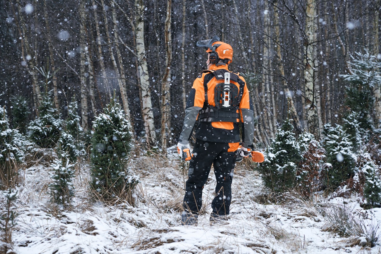 Lucrător forestier privit din spate, într-o pădure acoperită cu zăpadă, cu echipament de protecție STIHL cu sistem modular de curele și utilaje cu acumulator pentru îngrijirea arboretului tânăr