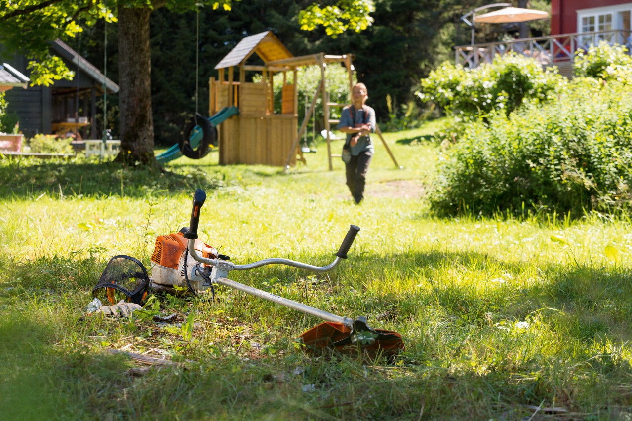 Motocoasa STIHL FS 240 se află în iarbă, iar pe fundal un bărbat se pregătește pentru întreținere