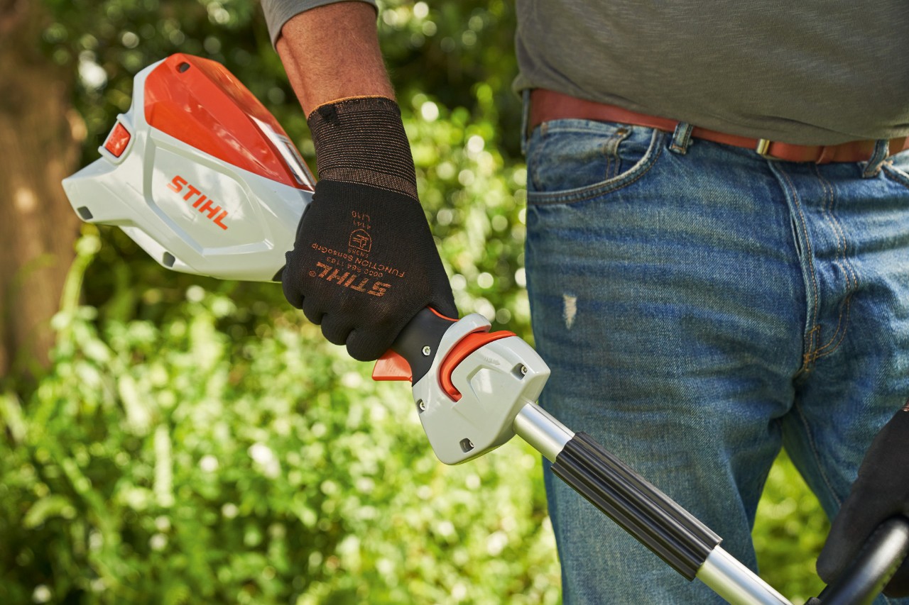 Un bărbat cu mănuși de protecție apasă blocatorul manetei de acționare a motocoasei cu acumulator STIHL FSA 60 