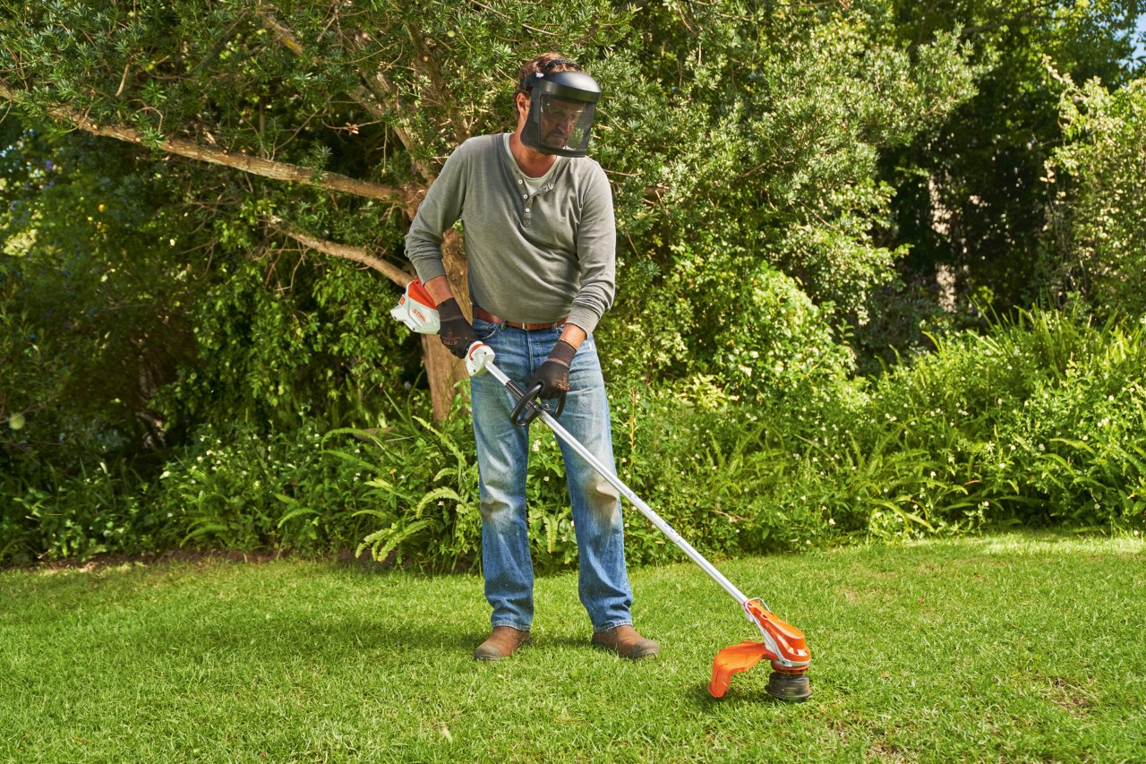 Un bărbat care poartă mască de protecție tunde gazonul într-o grădină cu motocoasa cu acumulator STIHL FSA 60