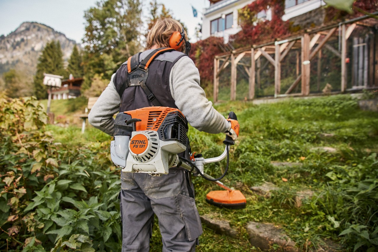 Iarba de pe terenul abrupt din fața unei case este tunsă cu motocoasa pe benzină STIHL FS 131 cu motor 4-Mix®